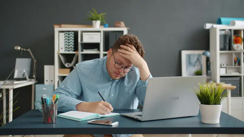 A man at an office desk with his hand on his forehead, writing notes next to a laptop.